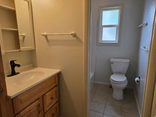 Dual-sink bathroom featuring wood-finish vanity with ample storage, ceramic tile flooring, and a separate water closet with natural light from a window