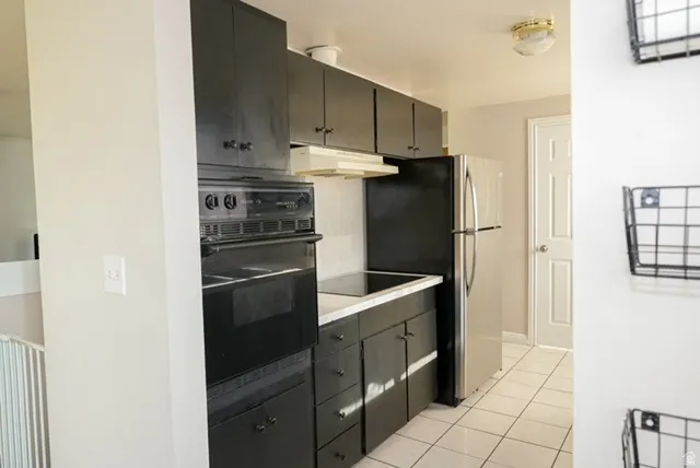 White tile flooring, built-in oven, black cabinetry, white countertops, and stainless steel refrigerator