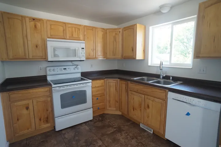 Kitchen featuring wood cabinetry, dark countertops, a double basin stainless steel sink, and a white appliance suite including an oven with cooktop, microwave, and dishwasher