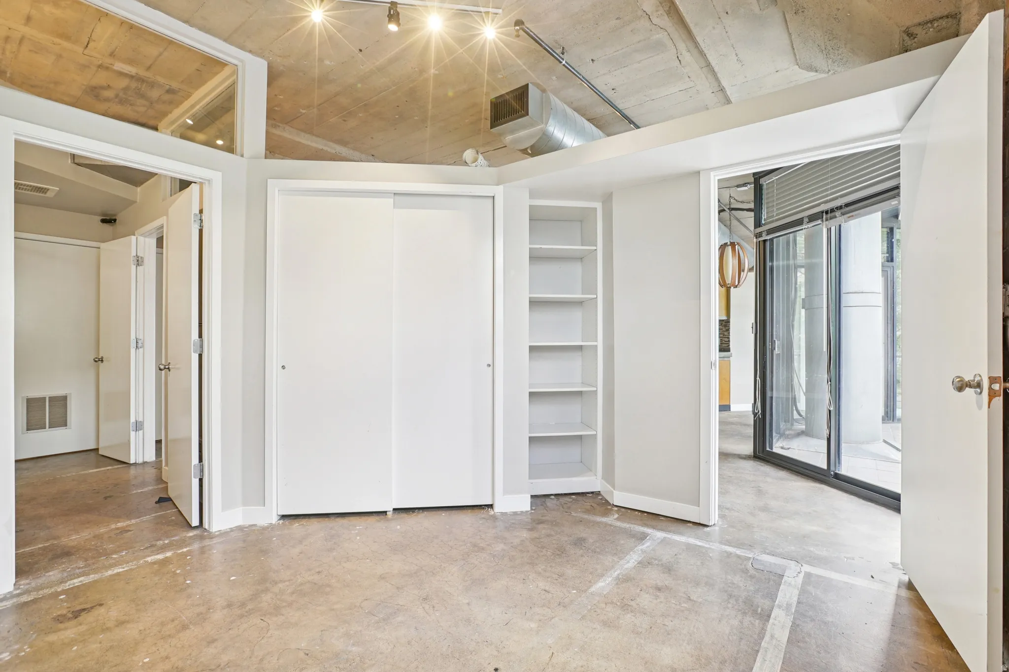 Exposed concrete ceiling with track lighting, polished concrete flooring, built-in shelving, white sliding closet doors, and a glass sliding door