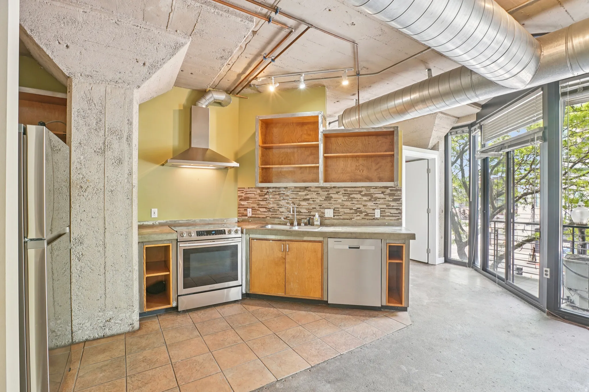 Exposed concrete ceiling with visible ductwork, stainless steel appliances, tile backsplash, built-in cabinetry, and full-height windows