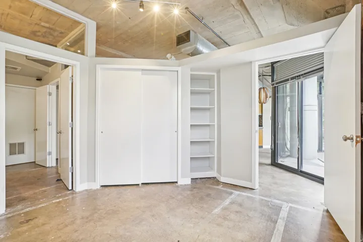 Exposed concrete ceiling with track lighting, polished concrete flooring, built-in shelving, white sliding closet doors, and a glass sliding door
