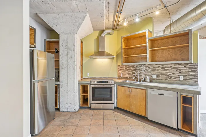 Stainless steel appliances, concrete countertops, mosaic tile backsplash, exposed concrete structural elements, and open shelving