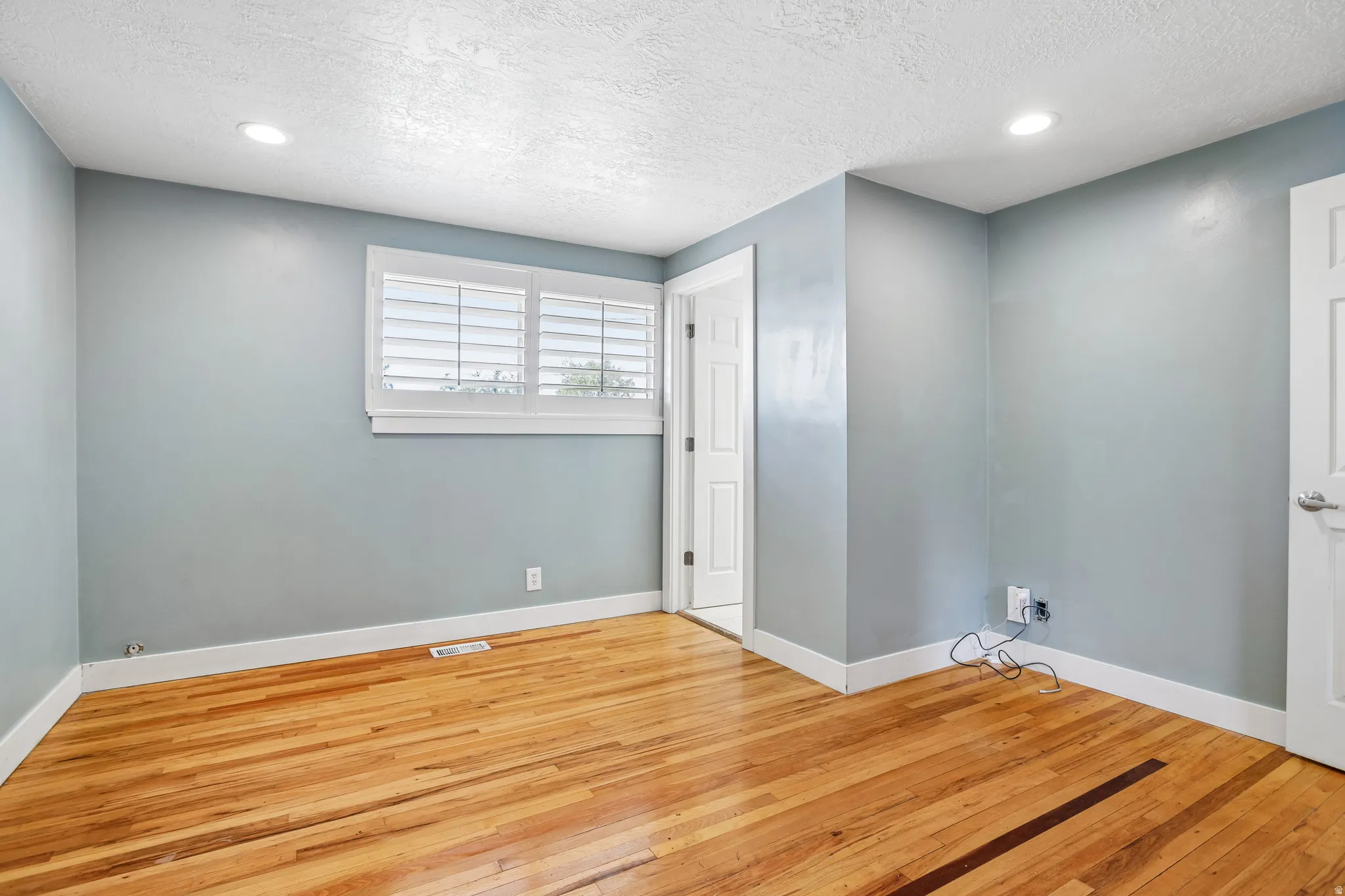 Hardwood flooring, recessed ceiling lighting, window with white plantation shutters, white trim, and light blue wall paint
