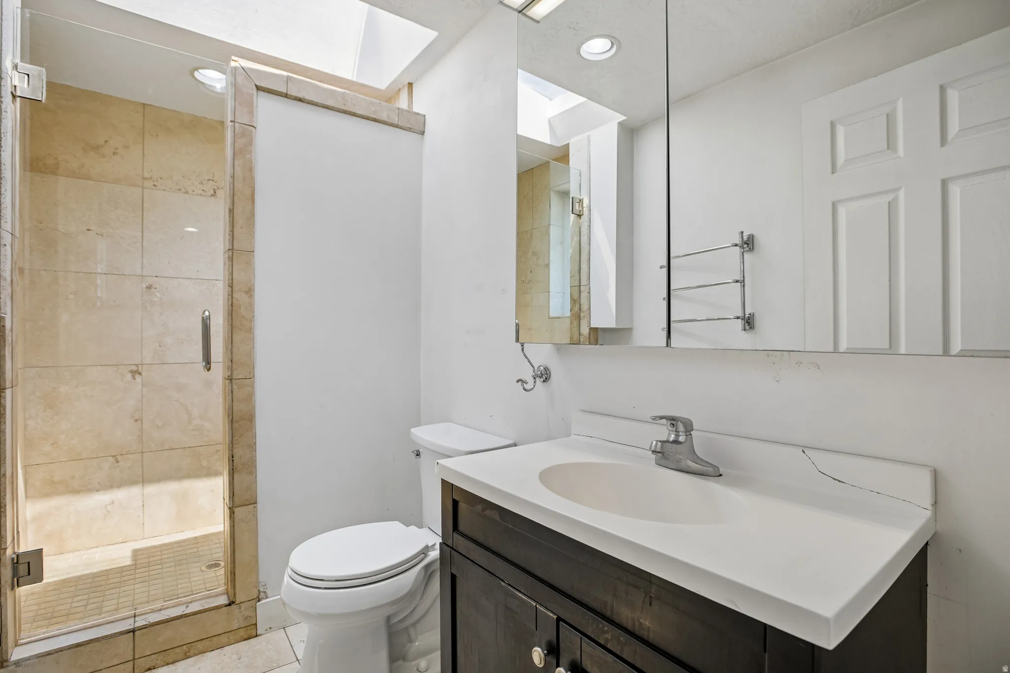 Glass-enclosed shower with stone tile surround and floor, wood-finish vanity with integrated sink, and overhead skylight