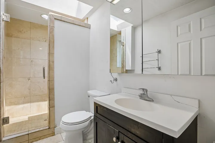 Glass-enclosed shower with stone tile surround and floor, wood-finish vanity with integrated sink, and overhead skylight