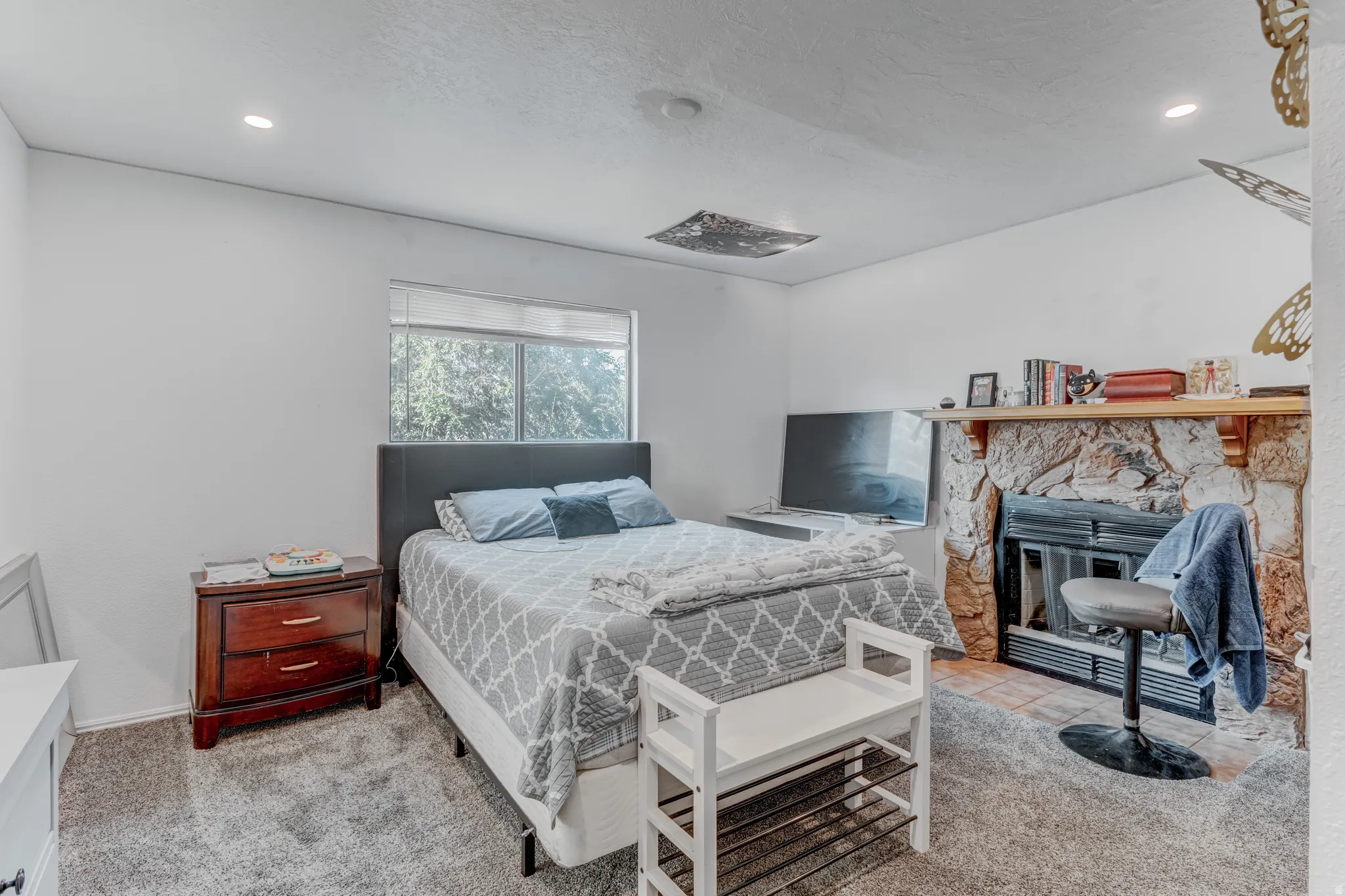 Recessed lighting, window with natural light, stone-clad fireplace with mantel, dark wood nightstand, and gray carpet flooring