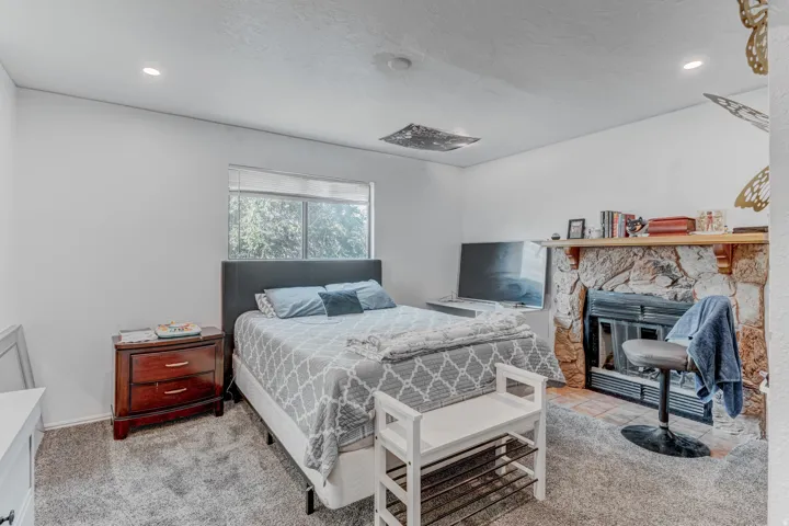 Recessed lighting, window with natural light, stone-clad fireplace with mantel, dark wood nightstand, and gray carpet flooring