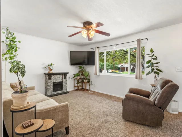 Carpeted living area featuring a ceiling fan, stone-finish electric fireplace, and a large window with white trim