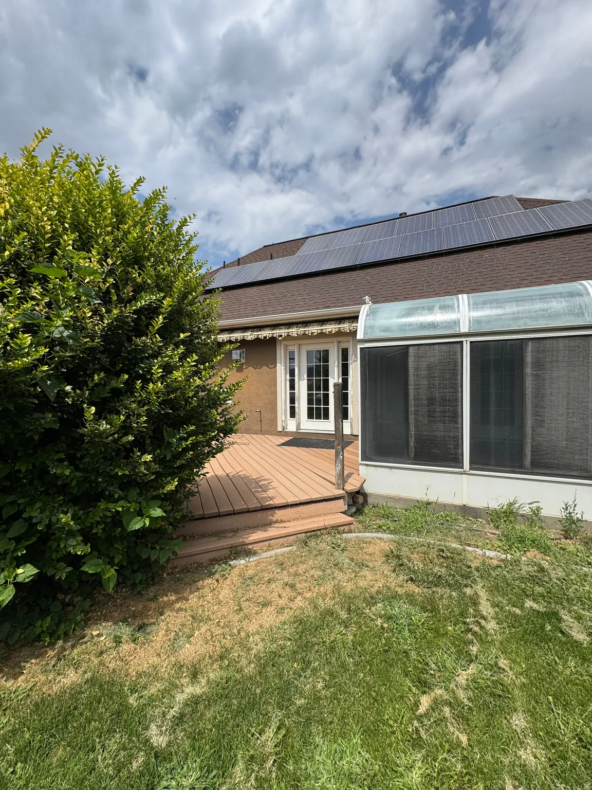 Brown shingle roof with solar panels, ground-level wood-finish deck, and greenhouse/sun room
