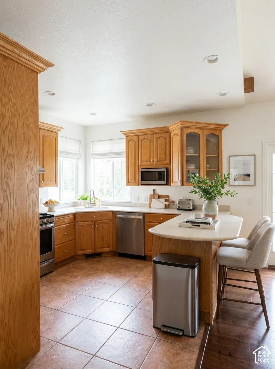 Recessed lighting illuminates the kitchen featuring wood-finish cabinetry, stainless steel appliances, a corner sink with a window, and a breakfast bar