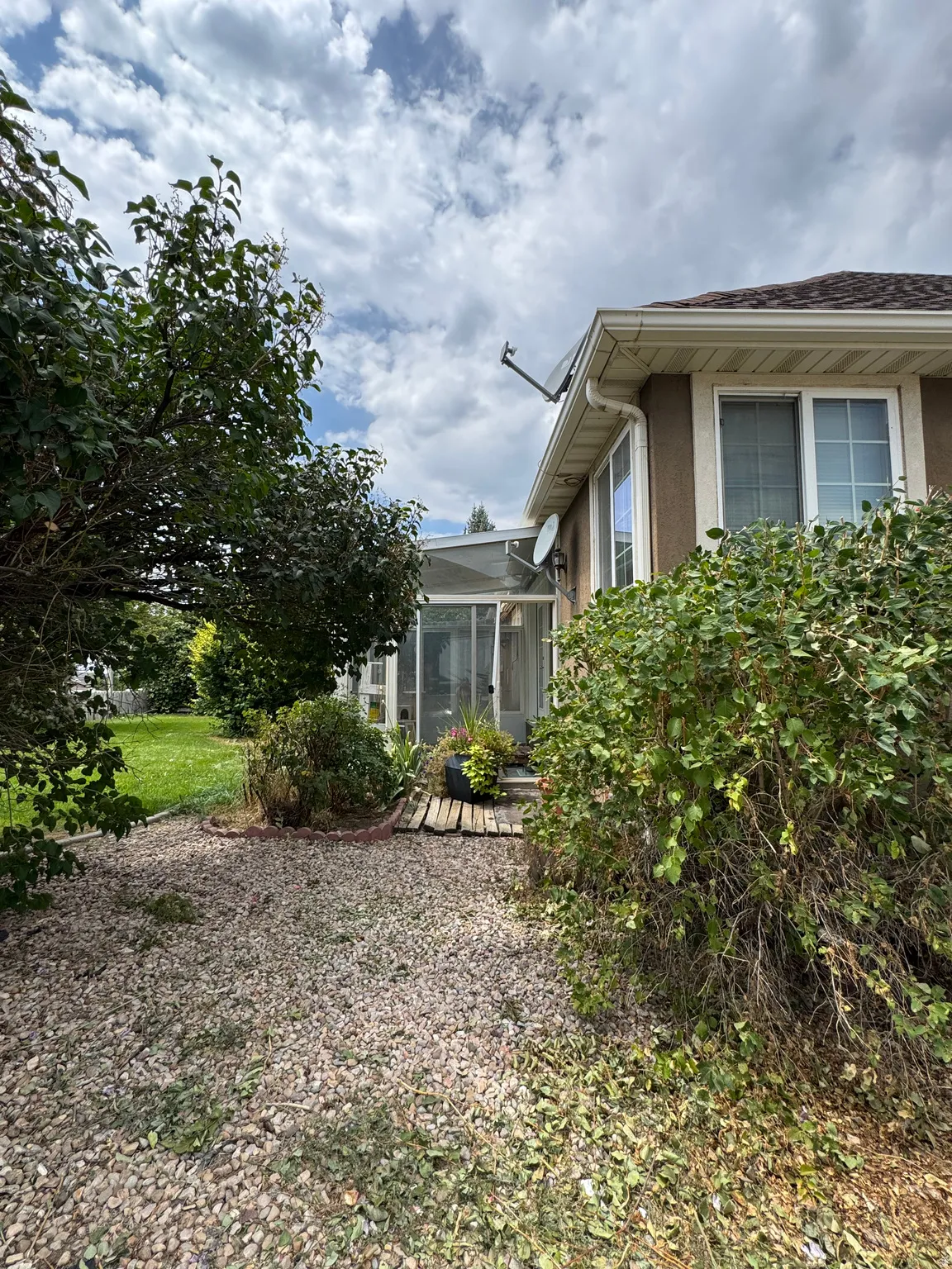Exterior wall with multiple windows, enclosed sunroom, gravel hardscaping, and established shrubbery