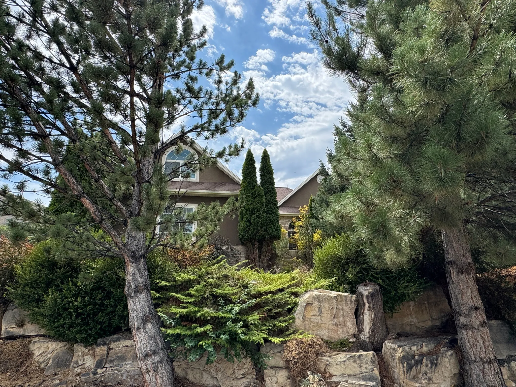 Exterior featuring an arched dormer window, established landscaping, and a natural stone retaining wall