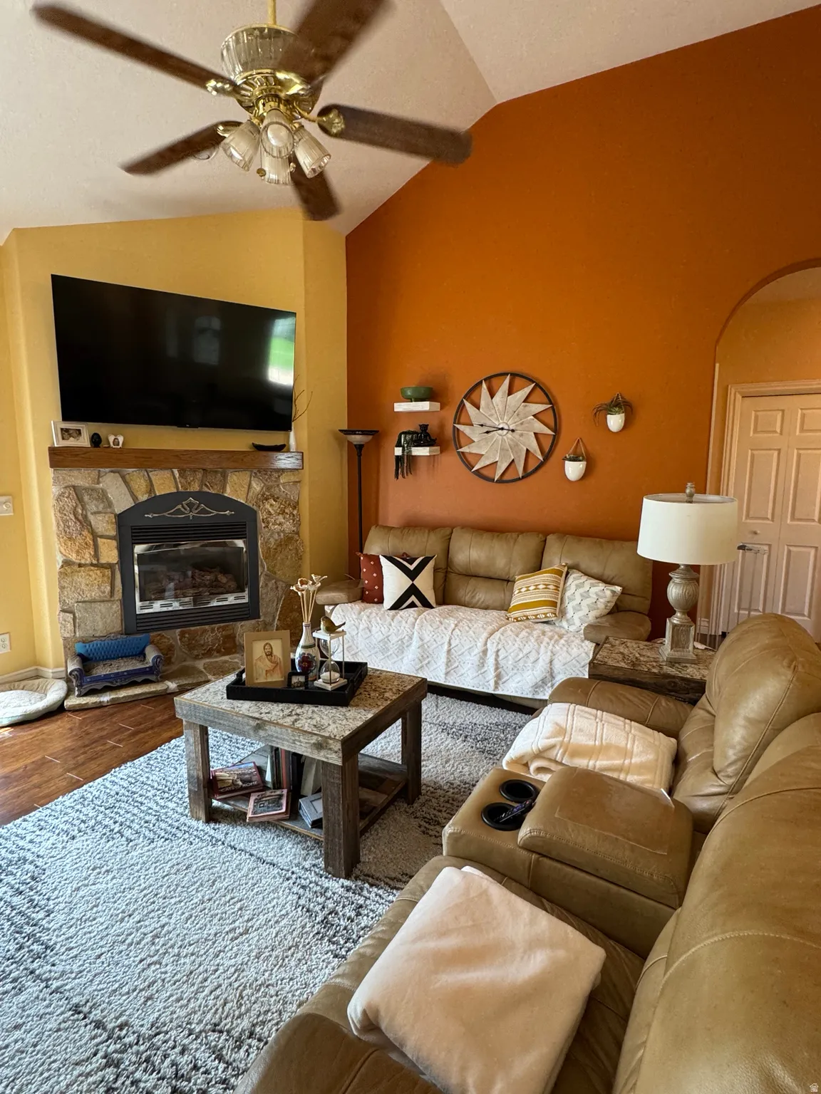 Vaulted ceiling with ceiling fan, stone-clad fireplace, wood-finish flooring, and recessed shelving