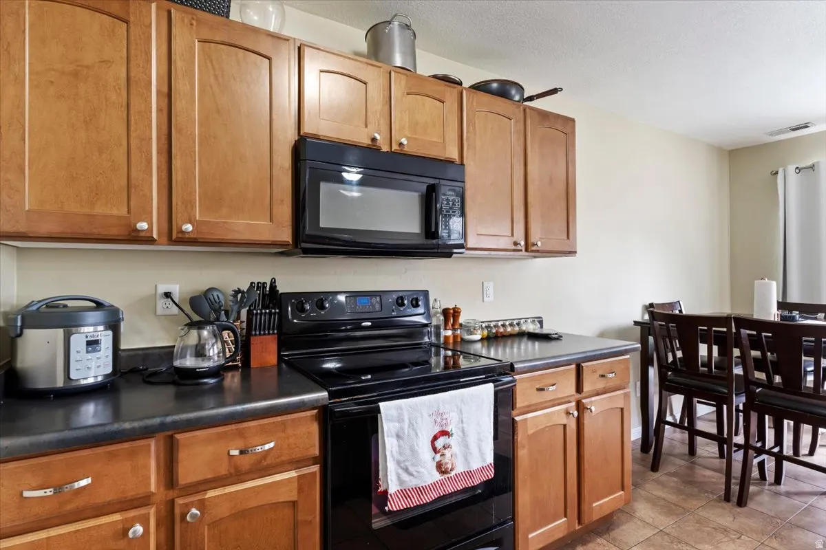 Wood-finish cabinetry with white hardware, black countertop, black over-the-range microwave, black electric range, and tiled flooring