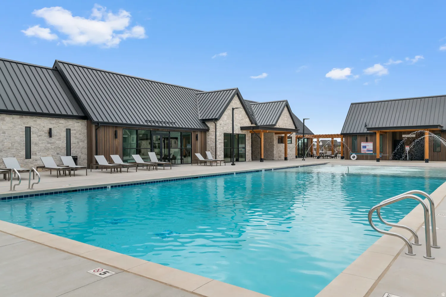 Community swimming pool with concrete decking, a water feature, stone and wood-finish siding buildings, and dark metal roofs