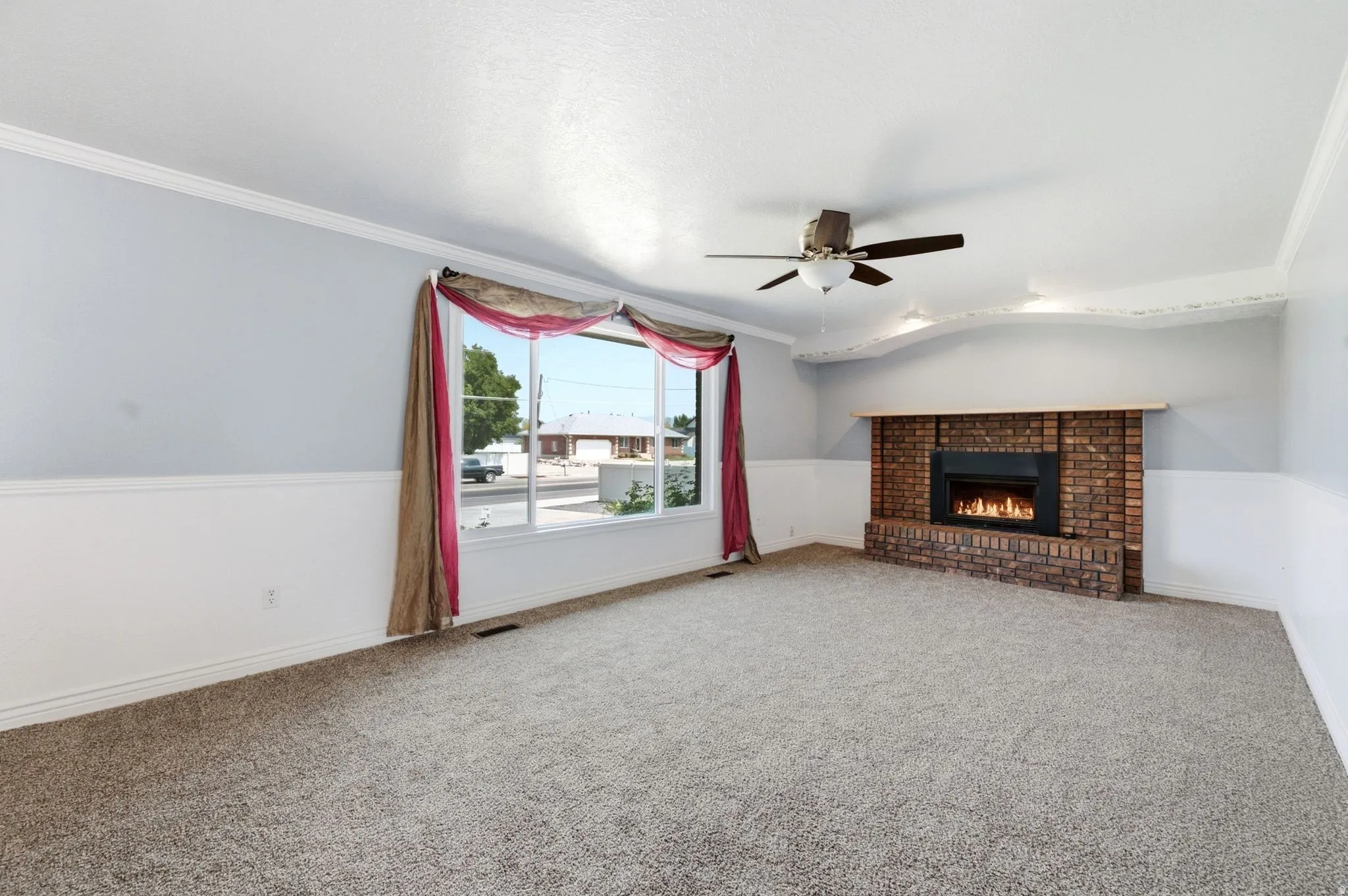 Brick-clad fireplace with a gas insert, ceiling fan, crown molding, chair rail molding, and a wide window