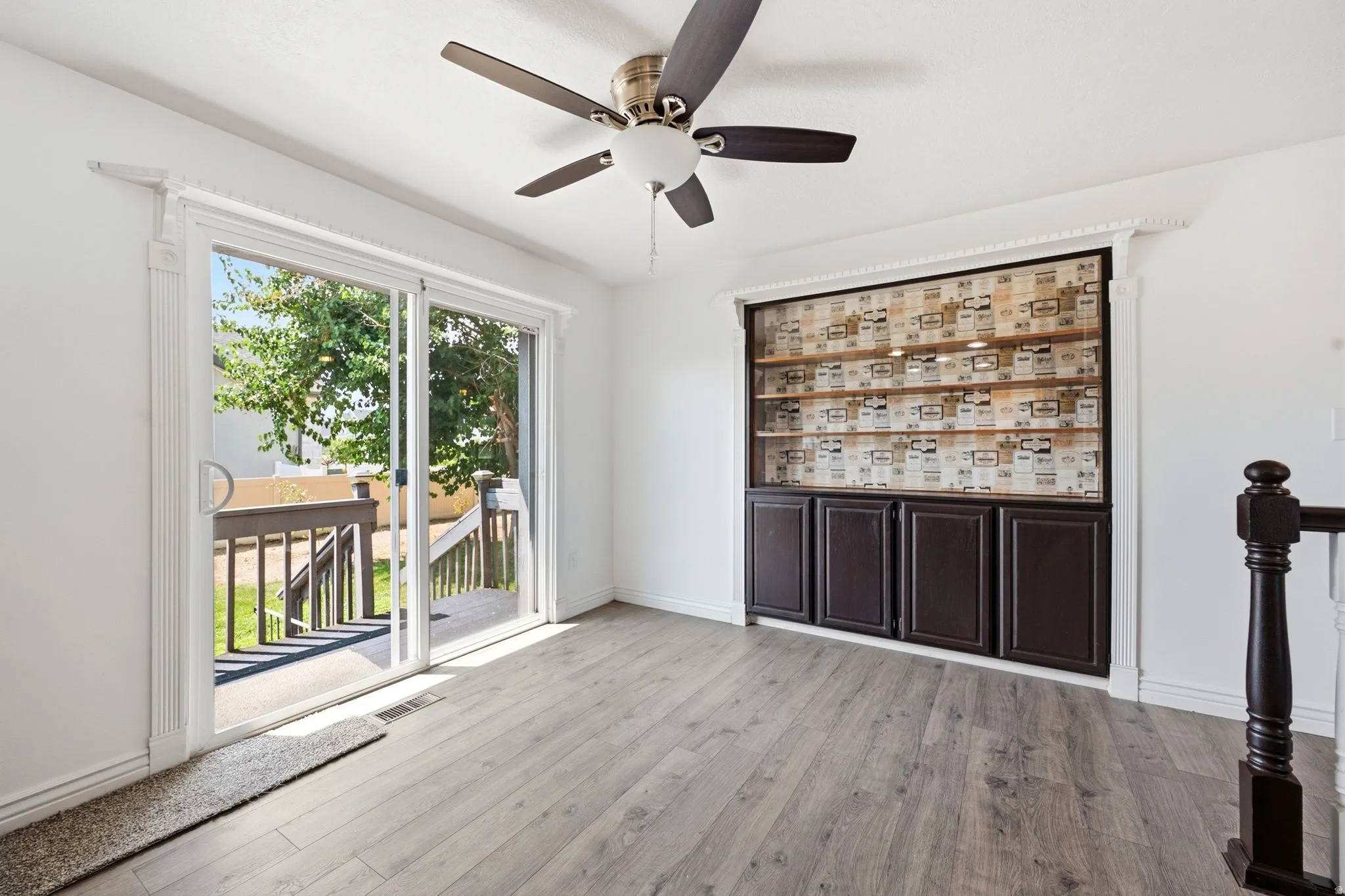 Sliding glass door to exterior deck, wood-finish flooring, built-in cabinetry with open shelving, ceiling fan with light fixture, and dark wood newel post