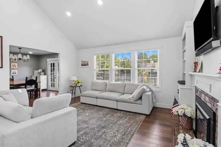 Vaulted ceiling with recessed lighting, wood-finish flooring, white trim windows, built-in shelving, and a fireplace with a white mantel and stone surround