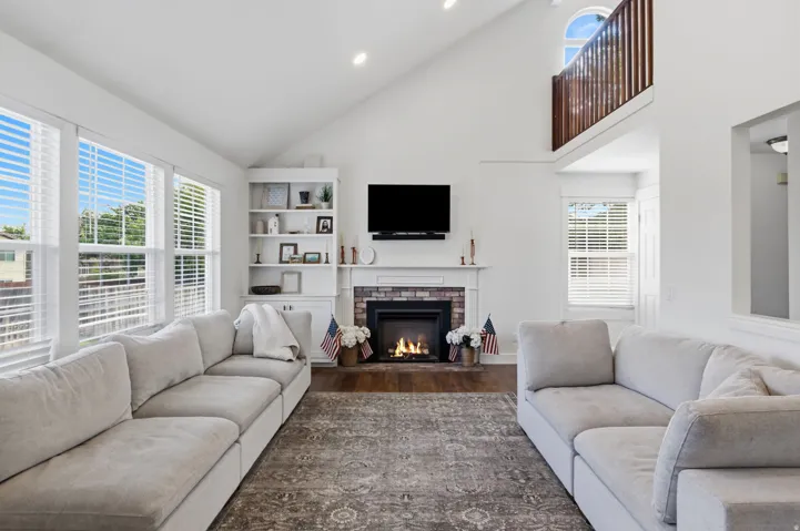 Soaring vaulted ceiling, wood-finish flooring, built-in shelving, fireplace with brick surround, and an upper-level arched window