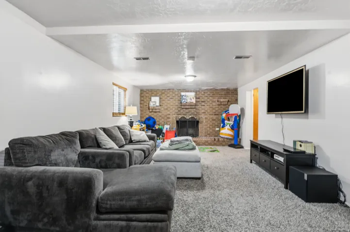 Spacious room featuring a brick fireplace, wall-mounted television, neutral carpeting, and a window with blinds