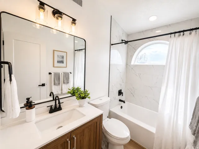 Bathroom featuring a wood-finish vanity with an integrated rectangular sink, a large framed mirror, a three-light fixture with clear glass shades, a tiled shower with a built-in tub, and an arched window
