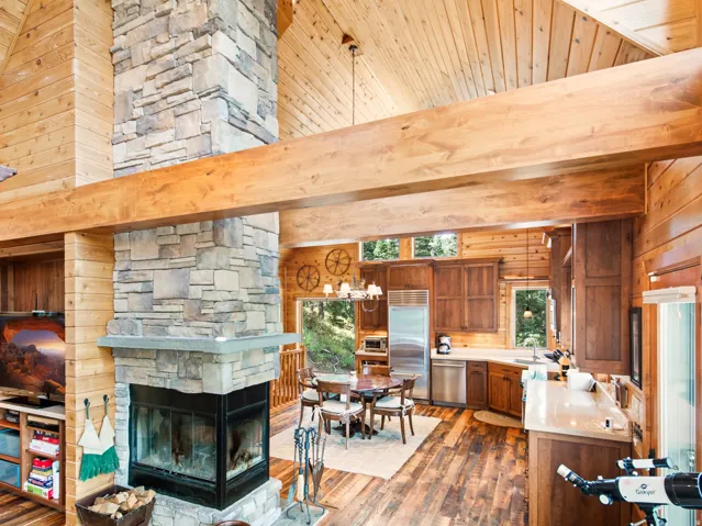 Vaulted ceiling with wood-finish planks, exposed wood beams, stone fireplace with hearth, built-in media cabinet, and a kitchen featuring wood cabinetry and stainless steel appliances