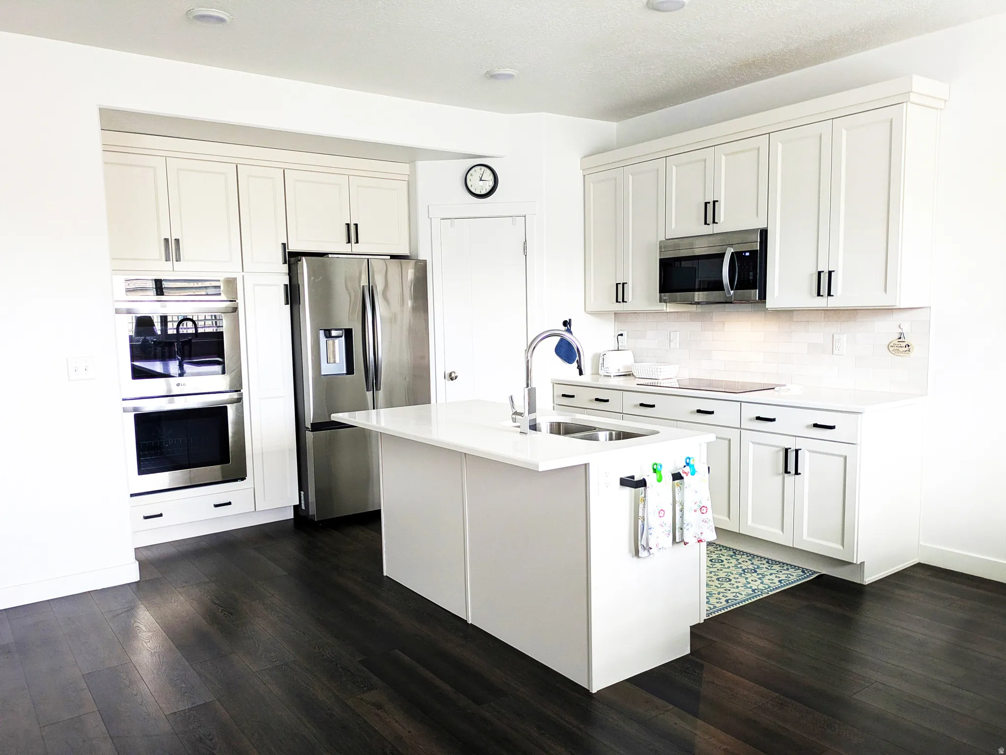 White shaker cabinetry, stainless steel appliances, dark wood-finish flooring, built-in double ovens, and a kitchen island with an integrated sink