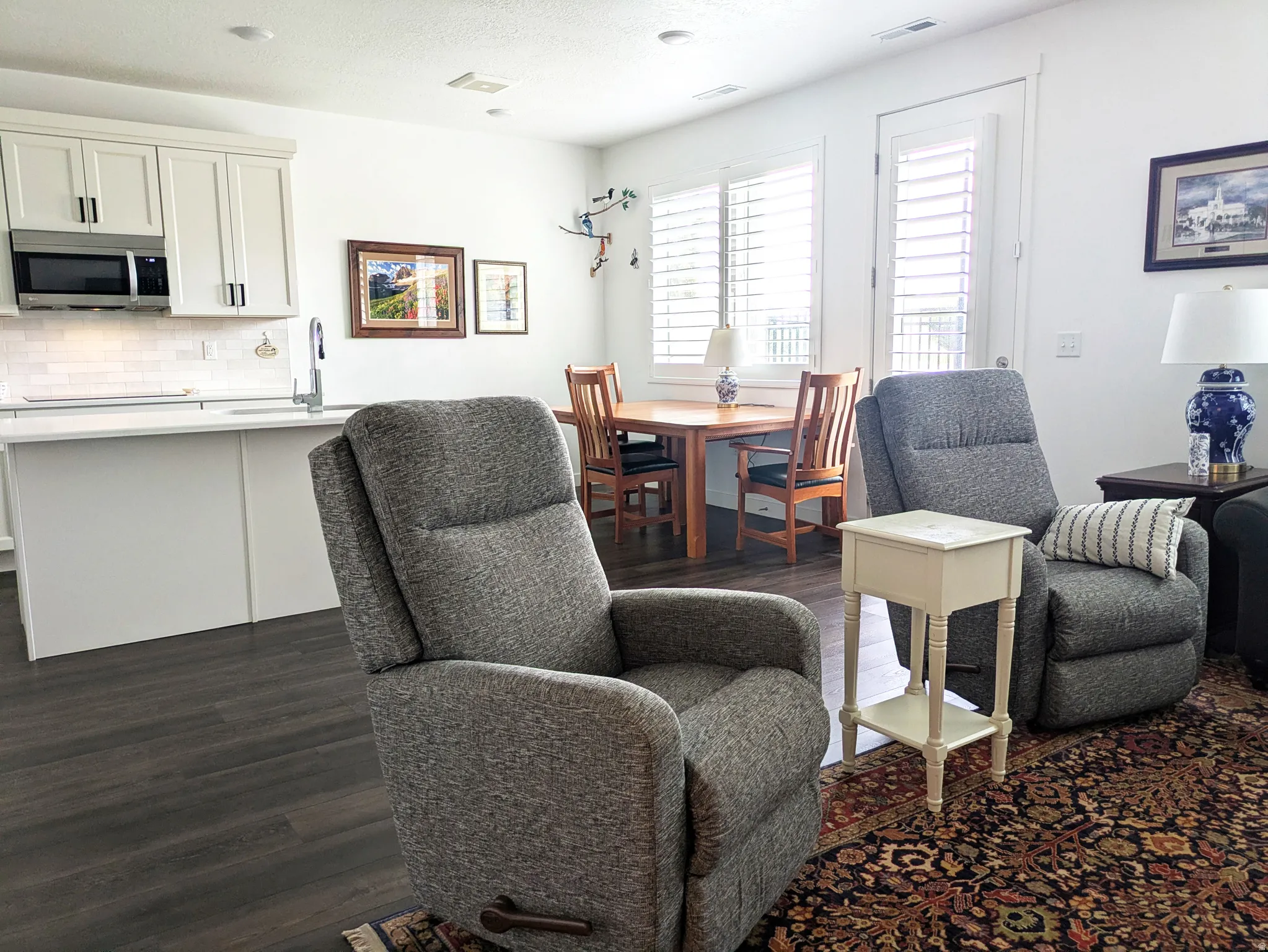 White shaker cabinetry, stainless steel microwave, white subway tile backsplash, dark wood-finish flooring, and white plantation shutters