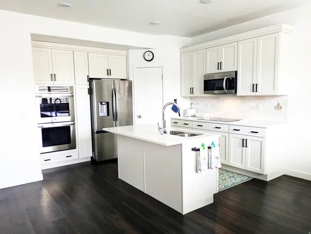White shaker cabinetry, stainless steel appliances, dark wood-finish flooring, built-in double ovens, and a kitchen island with an integrated sink