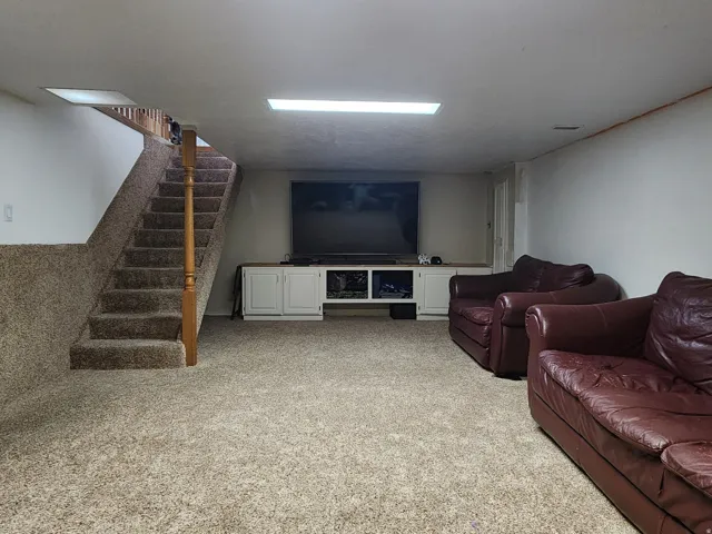 Spacious basement recreation area featuring neutral carpet flooring, recessed ceiling lighting, wooden staircase with railing, and built-in cabinetry with integrated entertainment storage