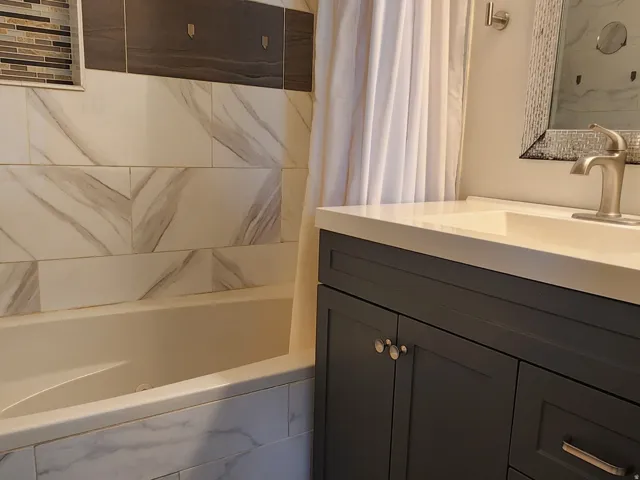 Bathtub with tiled surround featuring a decorative accent band and recessed niche, wood-finish flooring, dark grey vanity with a white countertop and integrated sink, and an ornate framed mirror