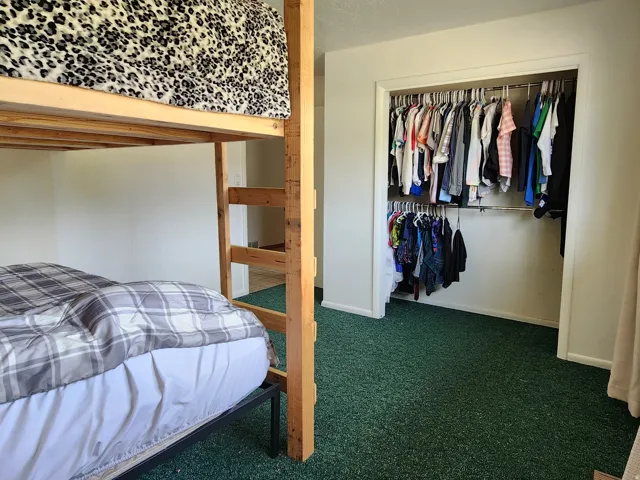 Built-in bunk bed frame, wall-to-wall green carpeting, open closet with two hanging rods, and a window with drapes