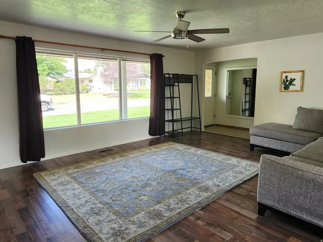 Wood-finish flooring throughout, ceiling fan with integrated lighting, large picture windows, and a built-in fireplace with a stone-finish hearth and white mantel