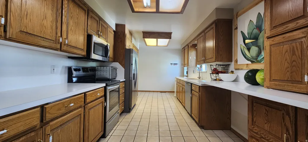 Galley kitchen featuring wood-finish cabinetry, stainless steel appliances, white countertops, tile flooring, and abundant natural light from skylights and ceiling fixtures