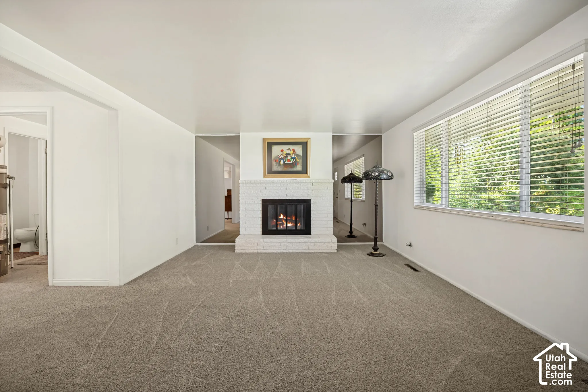 Carpeted interior space featuring a white brick fireplace with a black firebox, a large window with horizontal blinds, and neutral wall tones