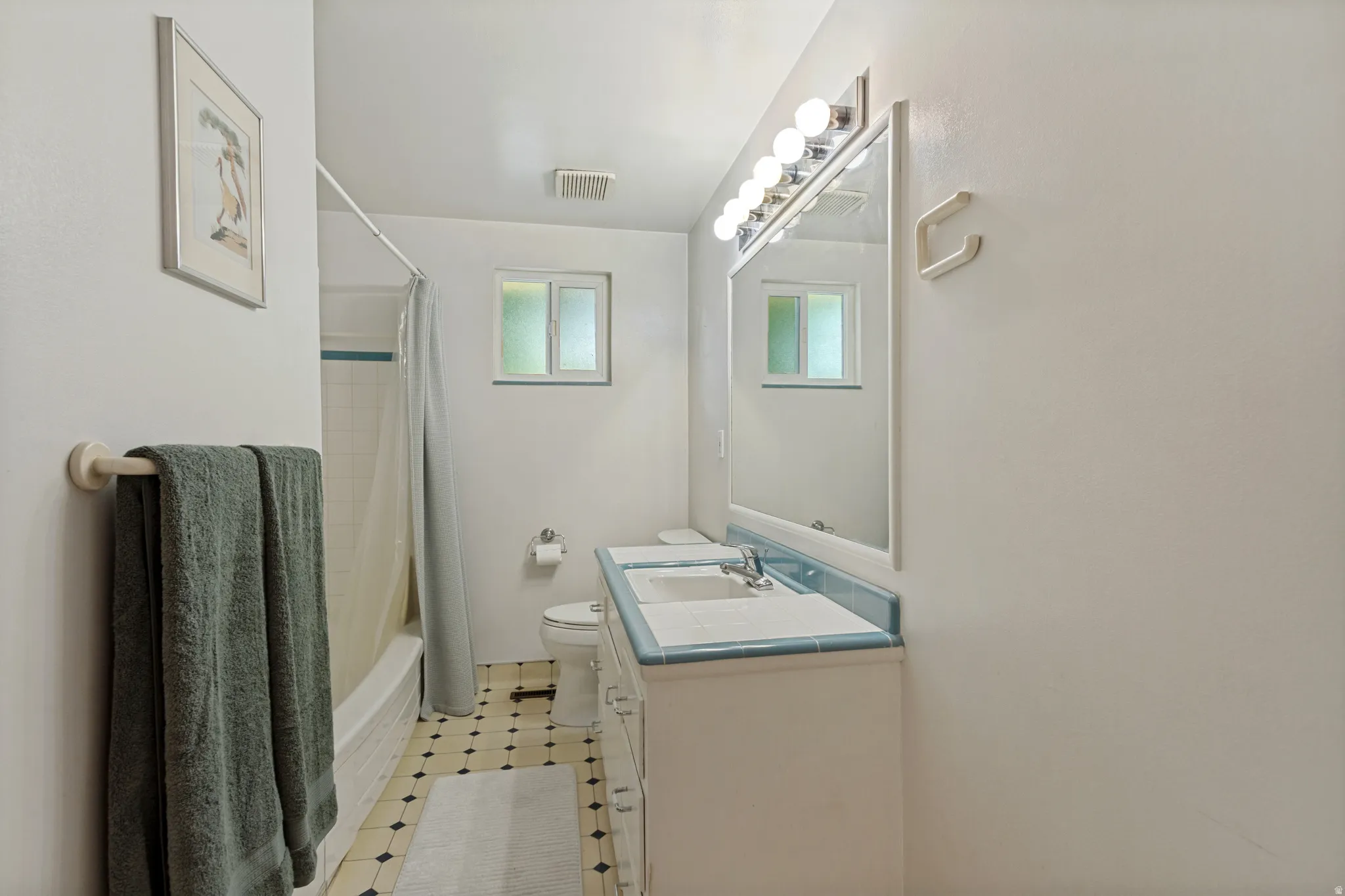 Bathroom featuring a built-in vanity with dual sinks, overhead vanity lighting, a large mirror, a window, and patterned floor tiling