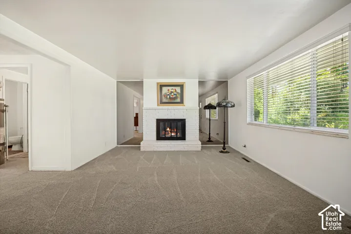 Carpeted interior space featuring a white brick fireplace with a black firebox, a large window with horizontal blinds, and neutral wall tones