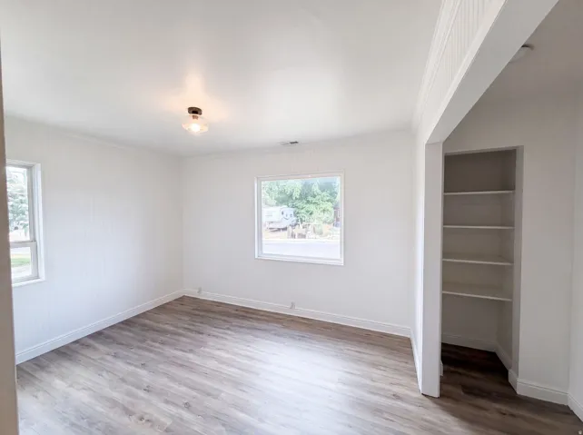 Spacious room featuring wood-finish flooring, two windows, wall paneling, and an integrated shelving unit