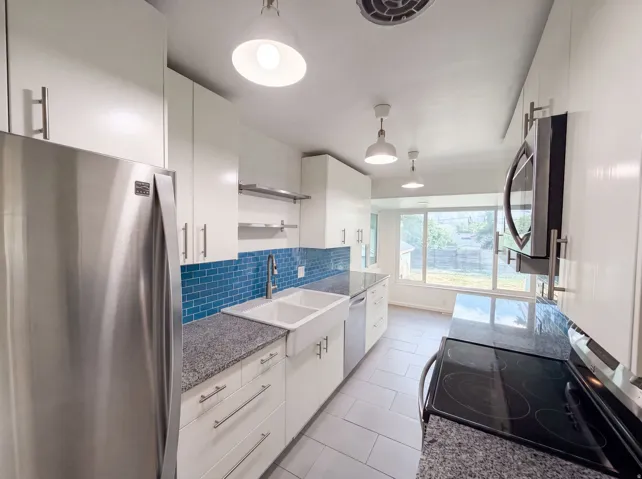 Modern kitchen with white cabinetry, stainless steel appliances, granite-look countertops, a blue tiled backsplash, and a double basin farmhouse sink