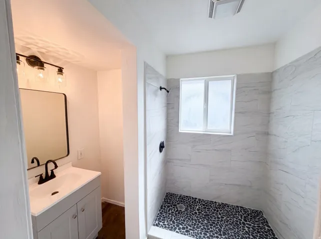 Vanity with a rectangular basin sink and matte black faucet, black-framed mirror, wood-finish flooring, tiled shower walls, and a pebble tile shower floor