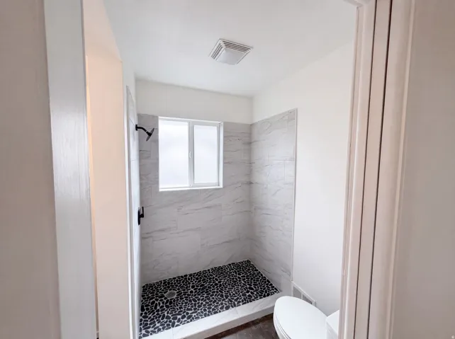 Walk-in shower with black pebble flooring, white tile surround, and a fixed window