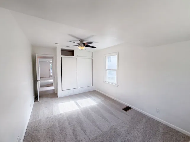 Grey carpet flooring, white painted walls, ceiling fan with integrated lighting, built-in storage with sliding doors, and a window with white trim