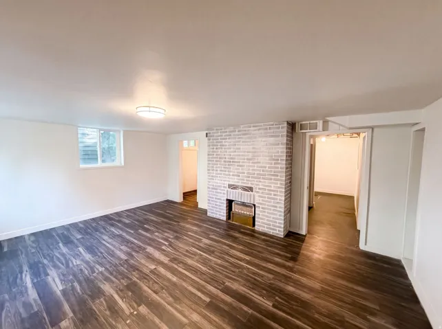 Spacious room featuring wood-finish flooring, a white brick fireplace, a ceiling-mounted light fixture, and a window with natural light