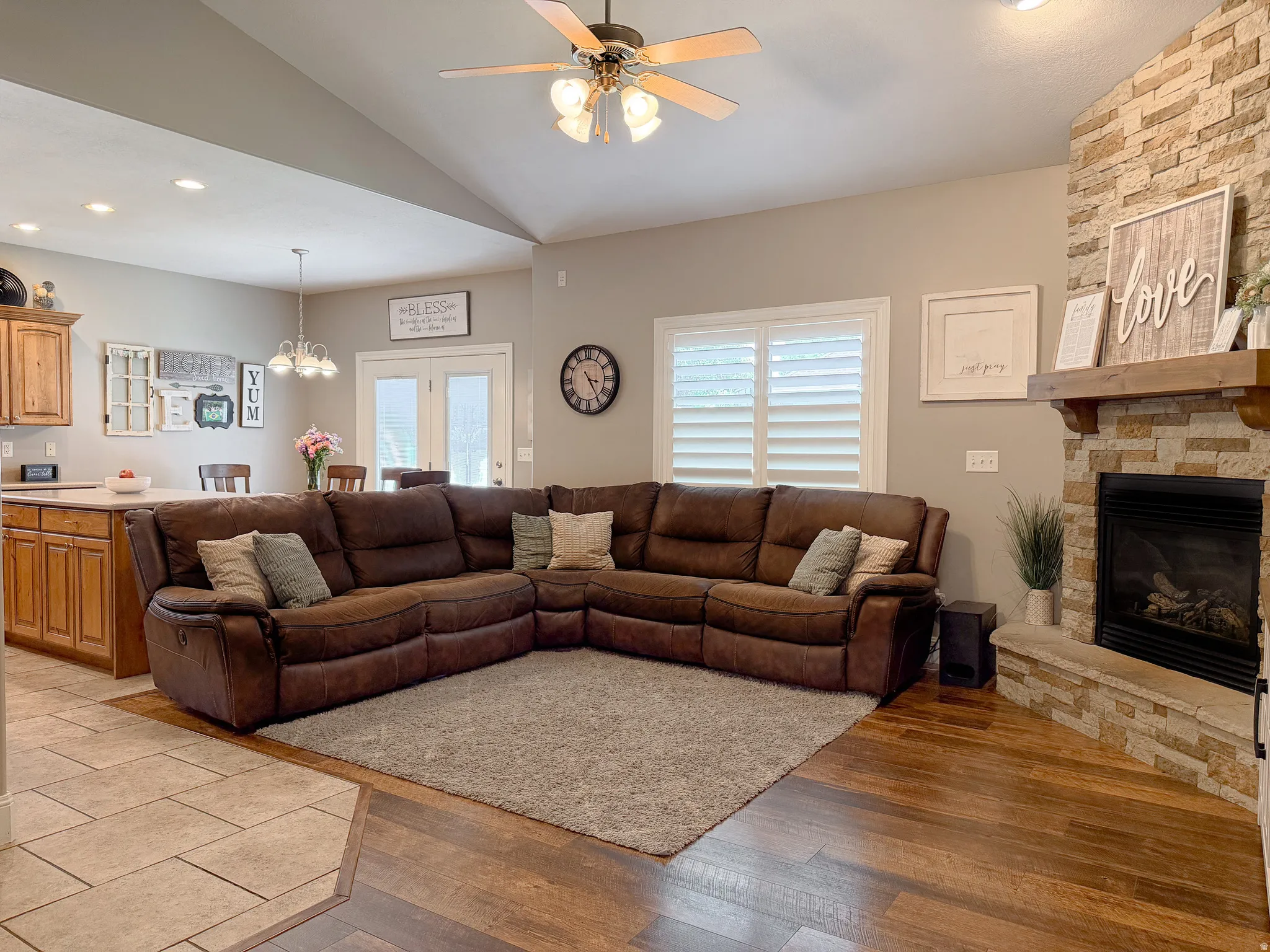 Vaulted ceiling with a ceiling fan, stone fireplace with a wood mantle, wood-finish flooring, white plantation shutters, and a recessed lighting
