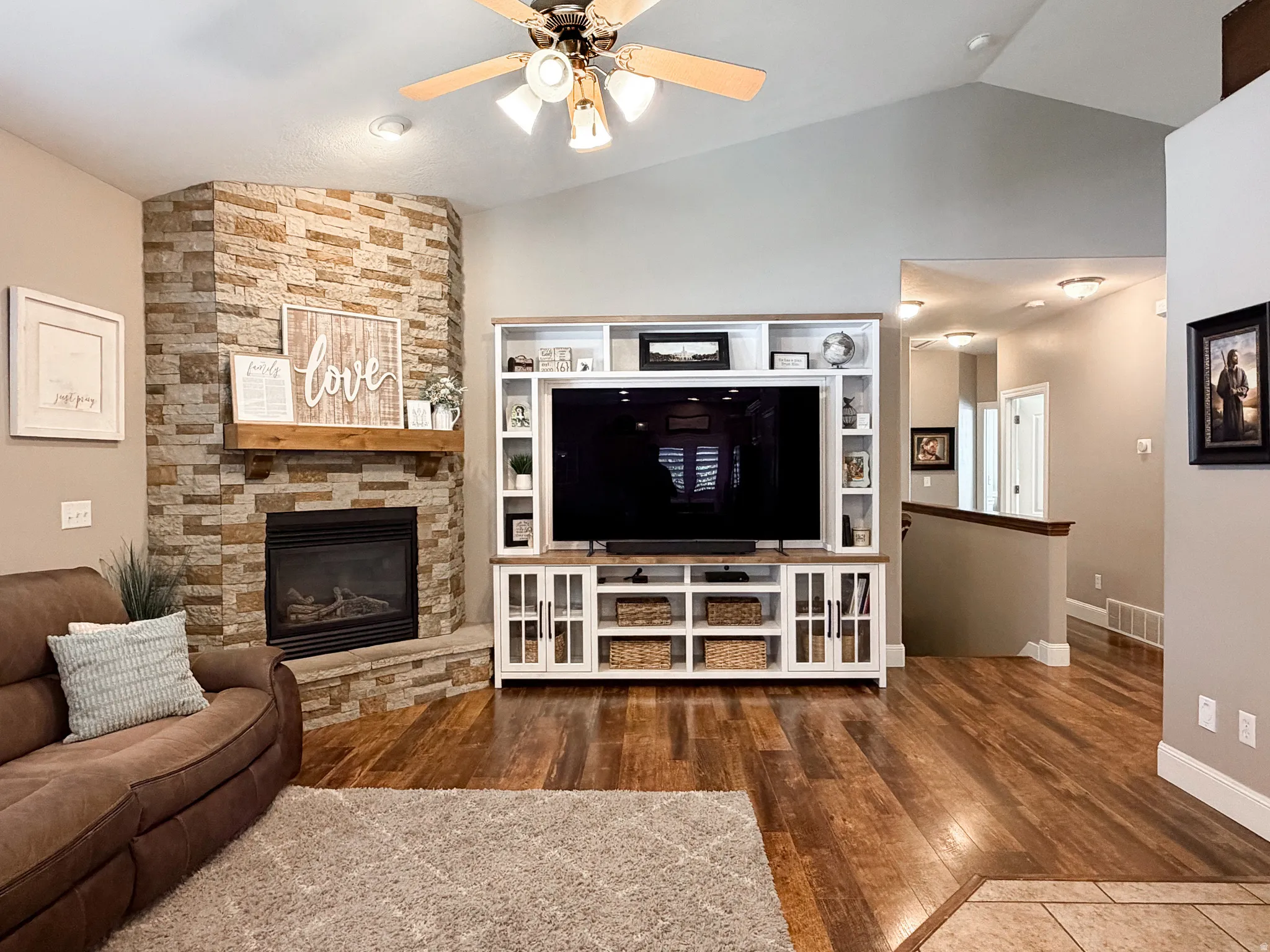 Corner stone-clad fireplace with wood mantle, built-in entertainment center with shelving, vaulted ceilings, wood-finish flooring, and recessed lighting