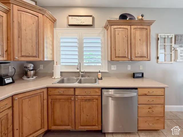 Wood-finish cabinetry with crown molding, double basin stainless steel sink with a gooseneck faucet, light-toned countertop, and a stainless steel dishwasher