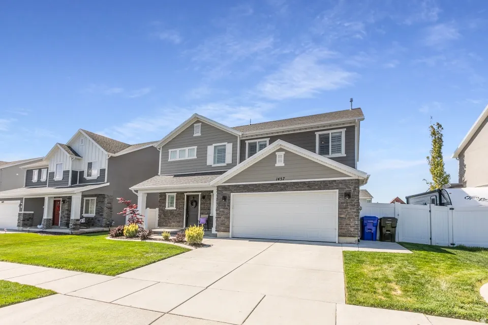 Two-story home featuring a stone and siding exterior, two-car attached garage with white overhead door, manicured front lawn, concrete driveway, and established landscaping with flowering plantings