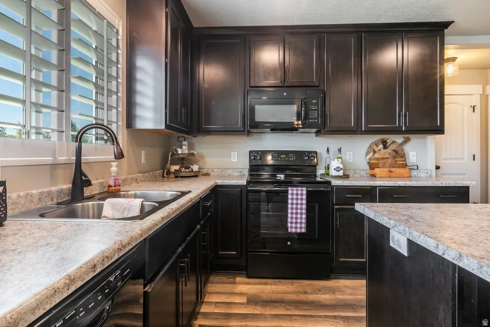 Kitchen featuring dark wood cabinetry, black appliances, light-toned countertops, and wood-finish flooring