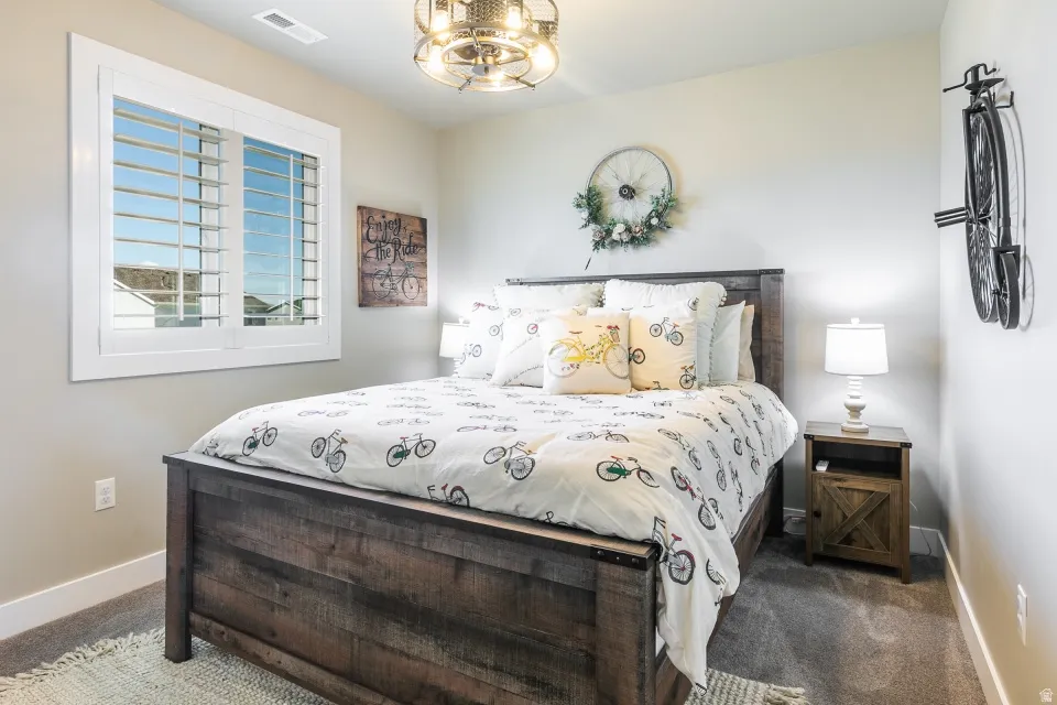 Wood-finish bed frame, carpeted flooring, window with white plantation shutters, ceiling-mounted light fixture, and light-toned wall paint